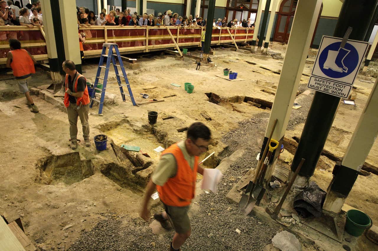 Archeologists wearing hi-vis vests go about their work on a small worksite with sections of dirt dug out from the ground, as people watch on from behind a barricade. 