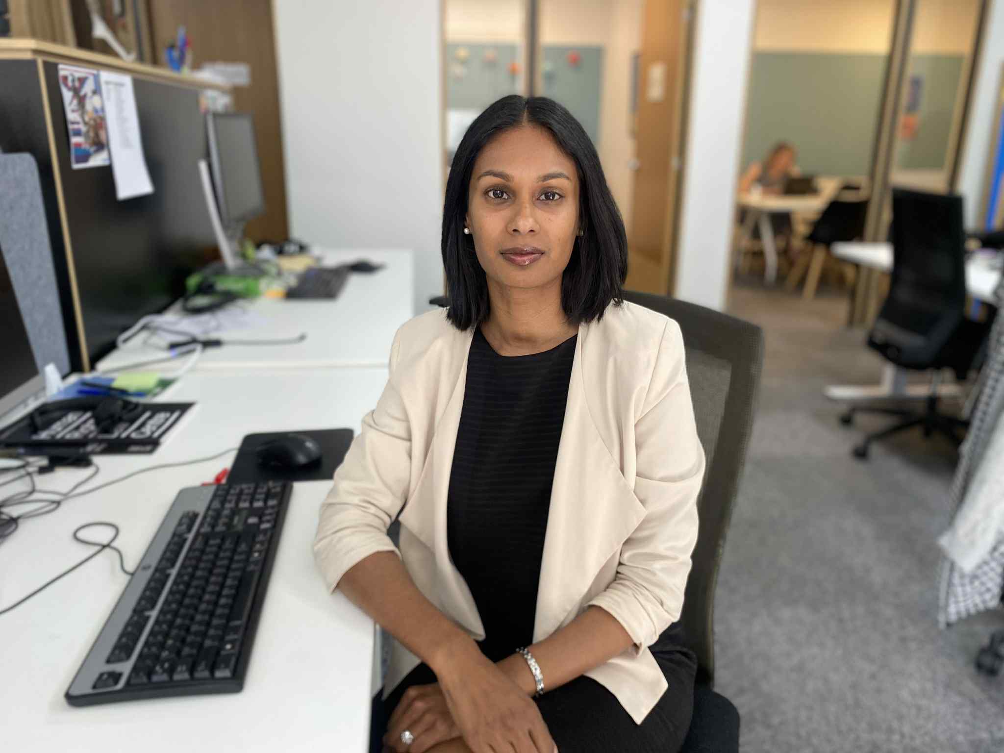 A woman with a dark straight bob, and brown skin, wearing a cream blazer and black top, sitting at a desk in front of a computer, in a typical open plan office setting.