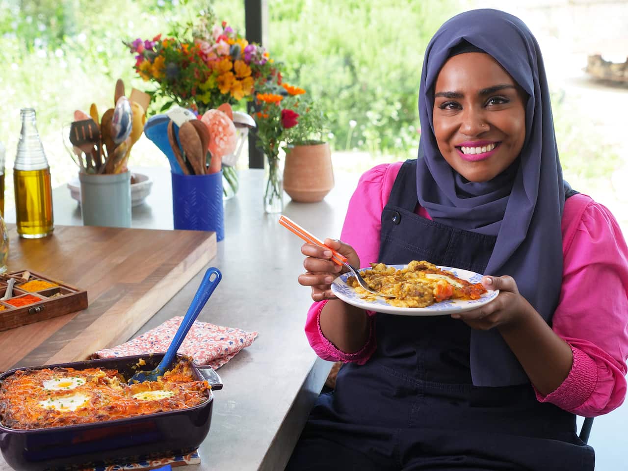 A smiling woman in a brik pink top with bue headscarf sits at a table. A baking tray of tray sits on the table, and she holds a serve of the dish in her hand. 