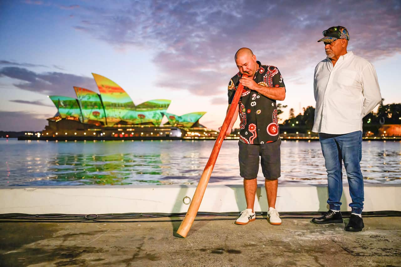 Two people, one holding a didgeridoo. The Sydney Opera House is lit up colourfully behind them.