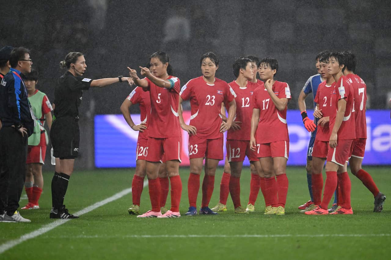 The North Korean women's team stand off towards the side of the pitch as it rains, as a player speaks to the referee while pointing to the distance with both hands