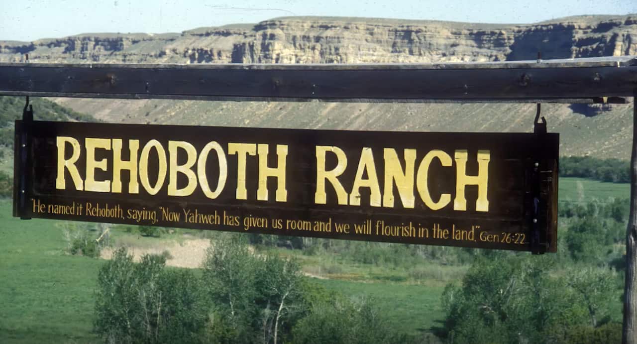A brown sign with yellow writing Rehoboth Ranch. In the background is a bare mountain range and in the foreground below, grassland.