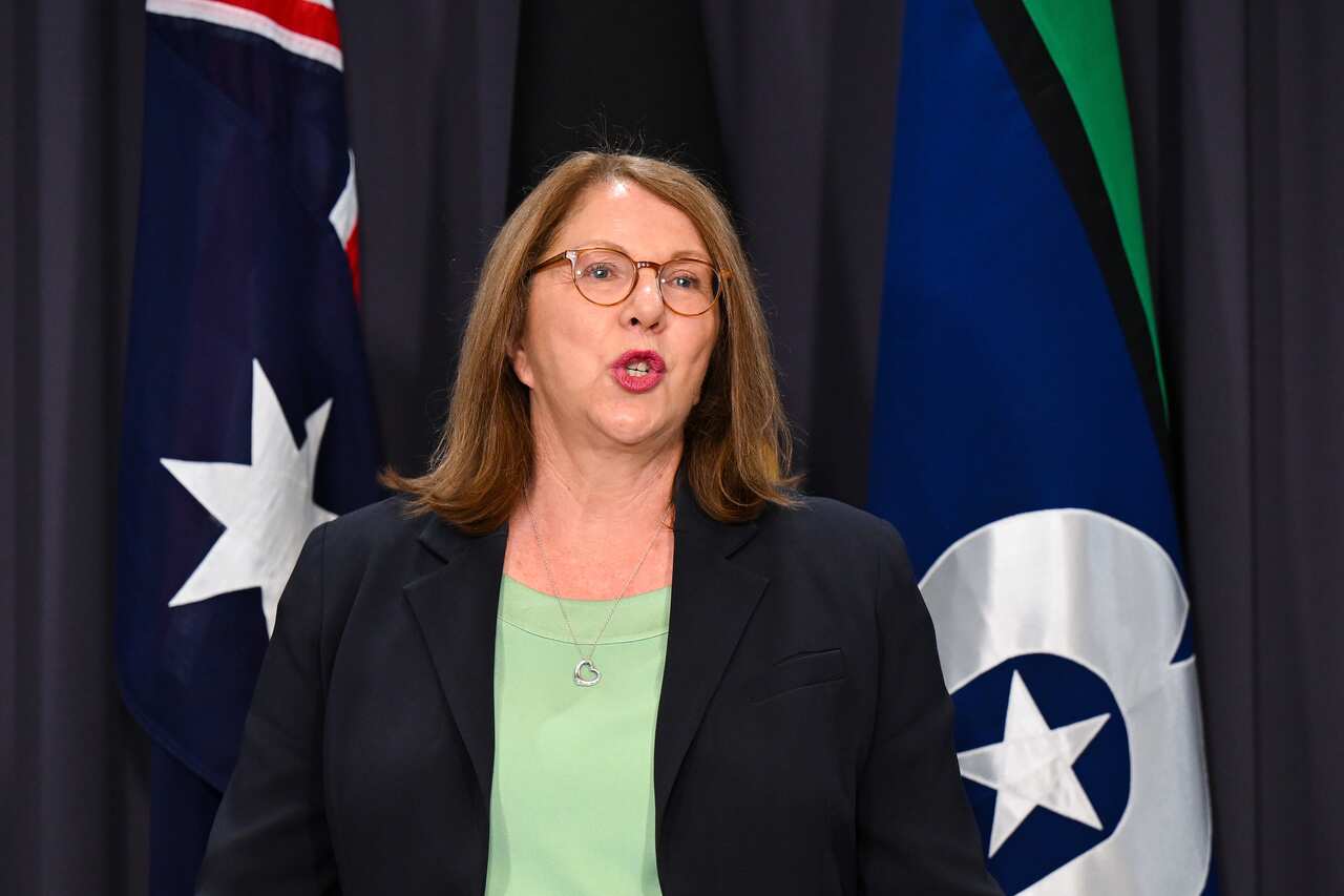 A woman in a black jacket standing and speaking in front of the Australian and Torres Strait Islands flags 