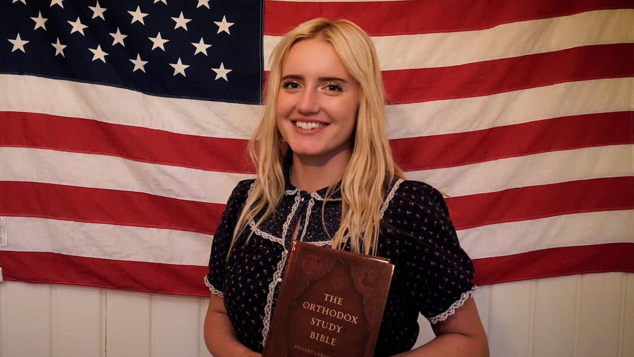 A blonde, smiling woman standings in front of an American flag while holding a book with the title 'The Orthodox Study Bible'. 