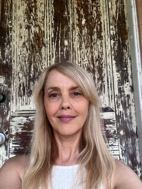 A woman with blonde hair and wearing a white sleeveless top poses for a selfie