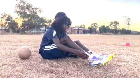 A teenage boy dressed in a black kit and pulling on football boots.
