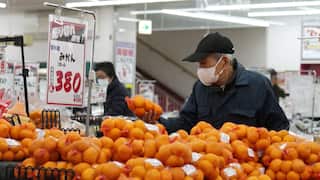 An elderly man wearing a dark baseball cap and jacket and a white medical mask is holding up a bag of potatoes at a supermarket