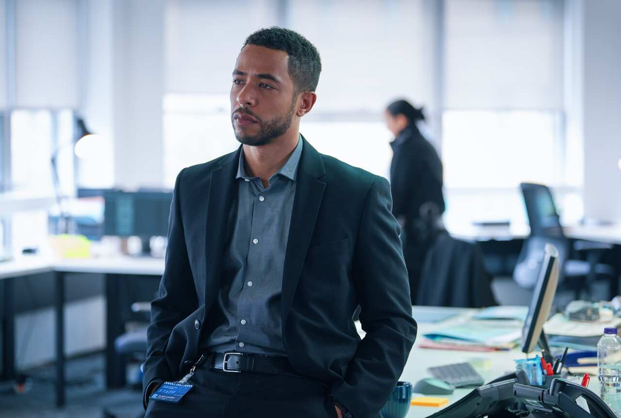 A man in a blue shirt and dark jacket leans on a desk in an office, looking pensively to one side.