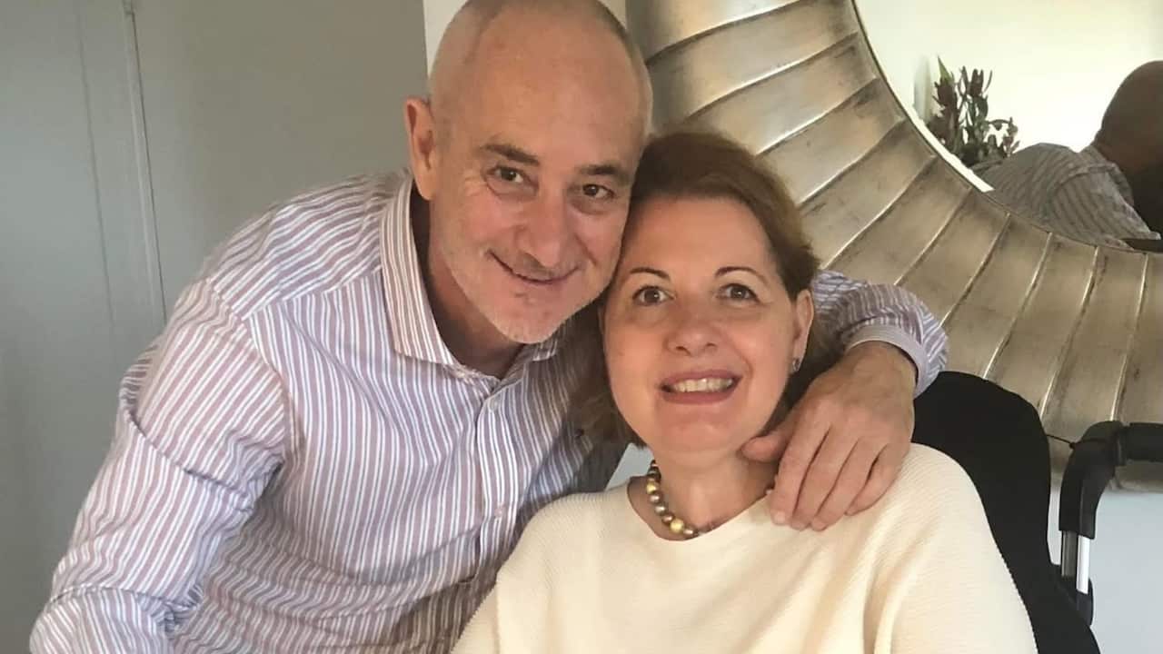 a closeup of a middle aged man and woman smiling. he has her arm around her and she is in a wheelchair