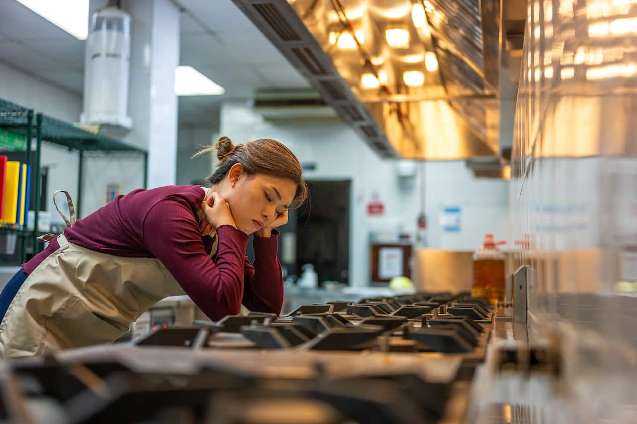 An Asian female chef looking at a tablet with an exasperated expression, resting her elbow on the kitchen counter and touching her forehead.