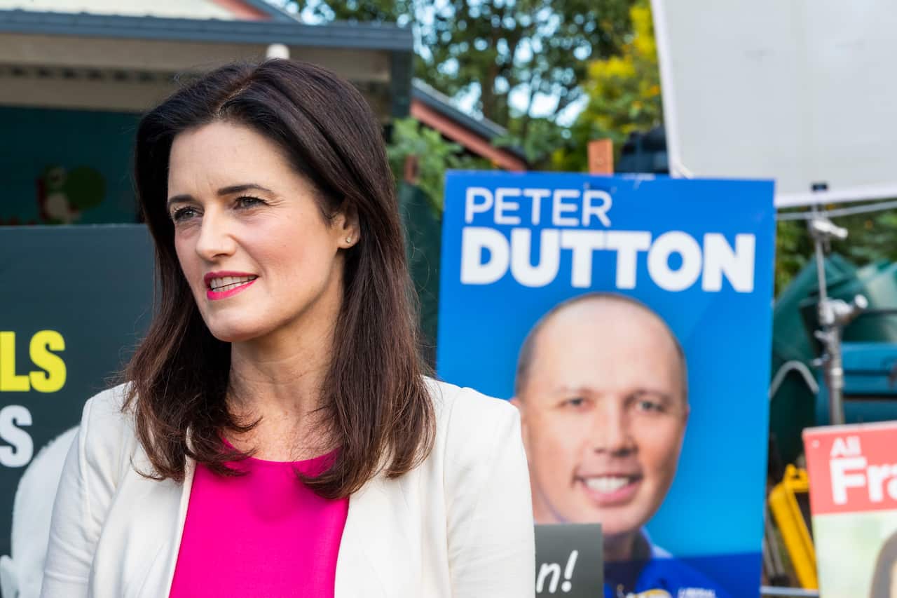 A brown haired woman with pink lipstick talking, with a poster of Peter Dutton in the background.