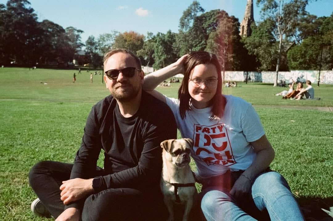 Two people smiling in a park. A small dog is sitting between them