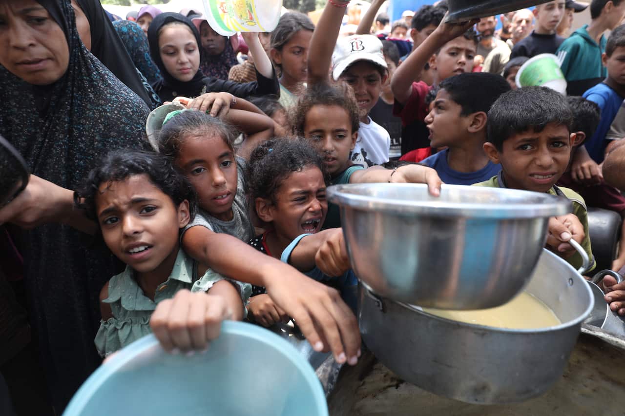 A group of people, including children, hold bowls to receive aid in a crushing crowd.