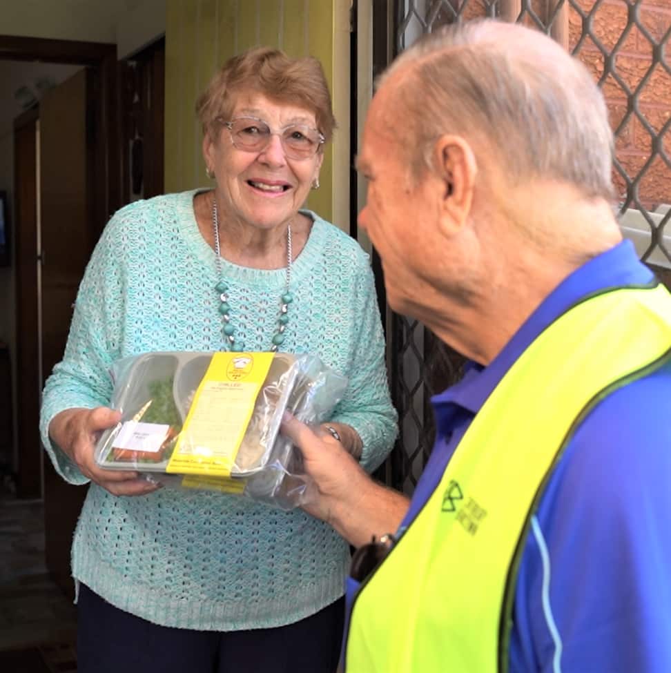 A man in a blue shirt and yellow hi-vis vest gives a meal to a woman at her front door. She is wearing glasses and a light green jumper.