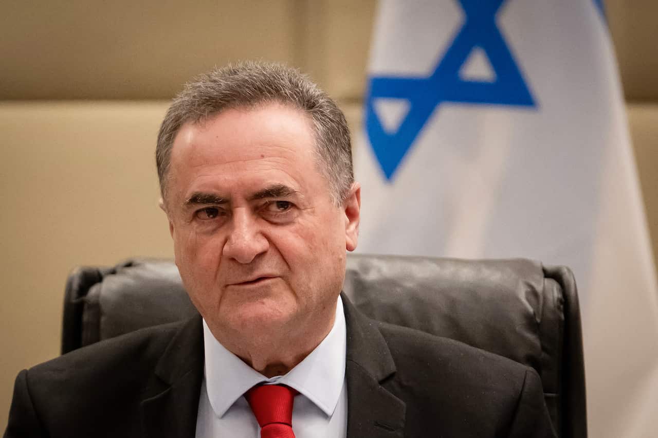 A man in a suit and red tie sits in a chair, with a serious expression, in front of an Israeli flag.