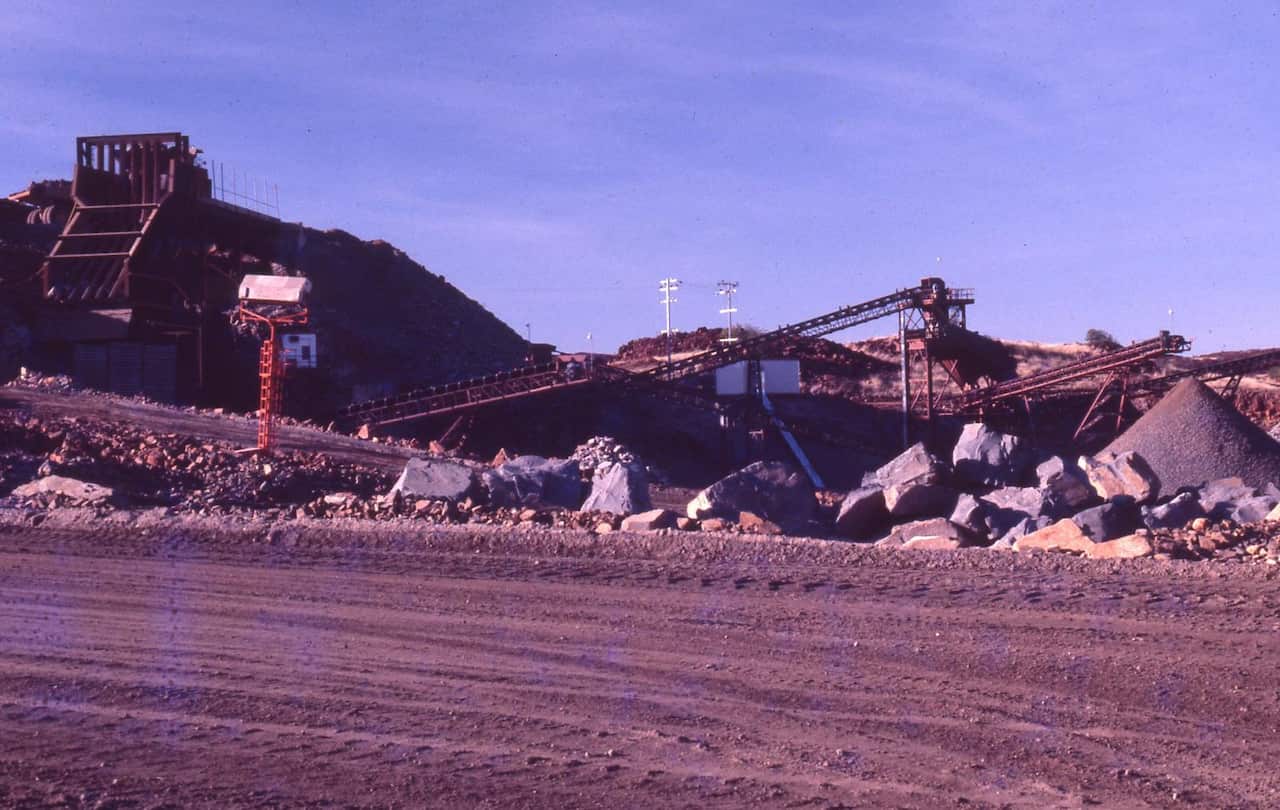 An industrial mining or quarrying site featuring heavy metal structures, conveyor belts, large piles of rocks.