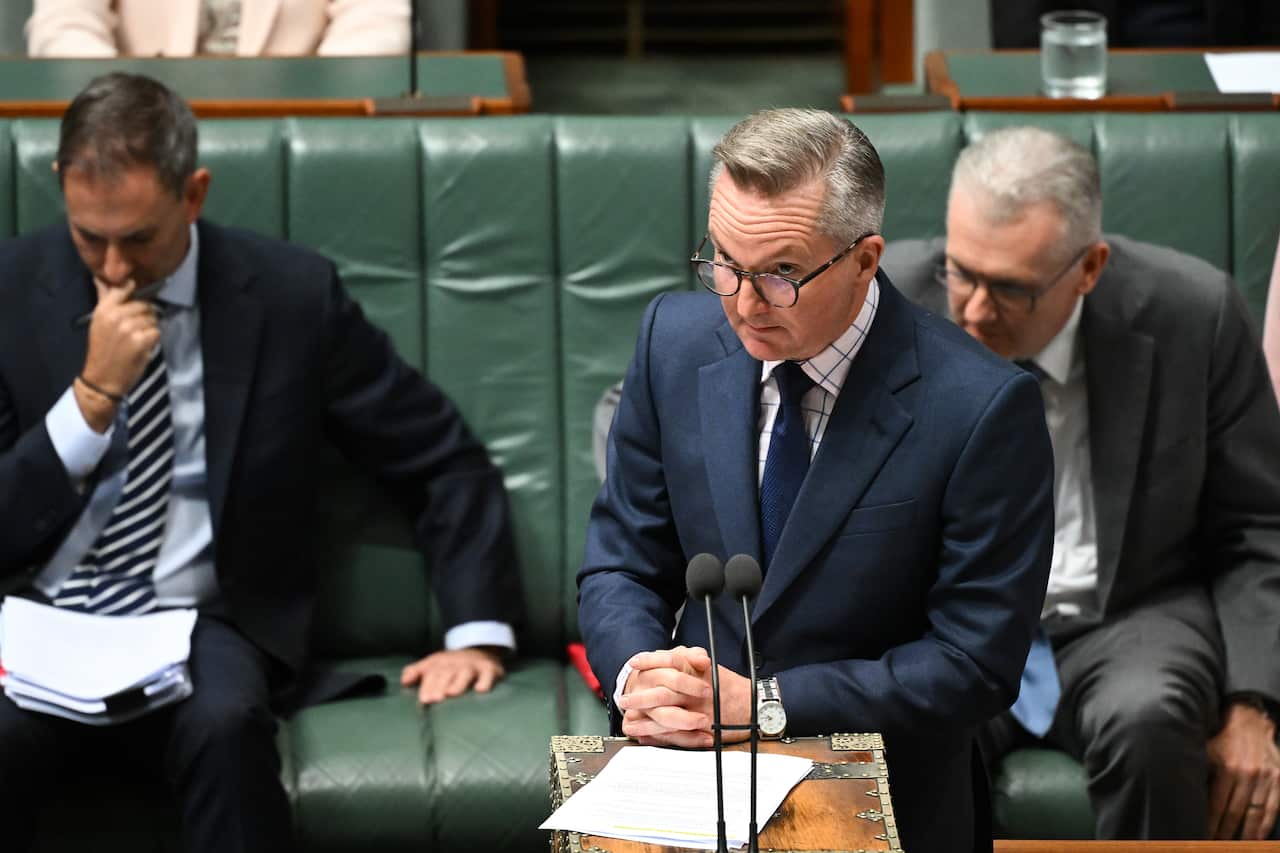 A man stands at a lectern in parliament, looking sternly over his glasses.