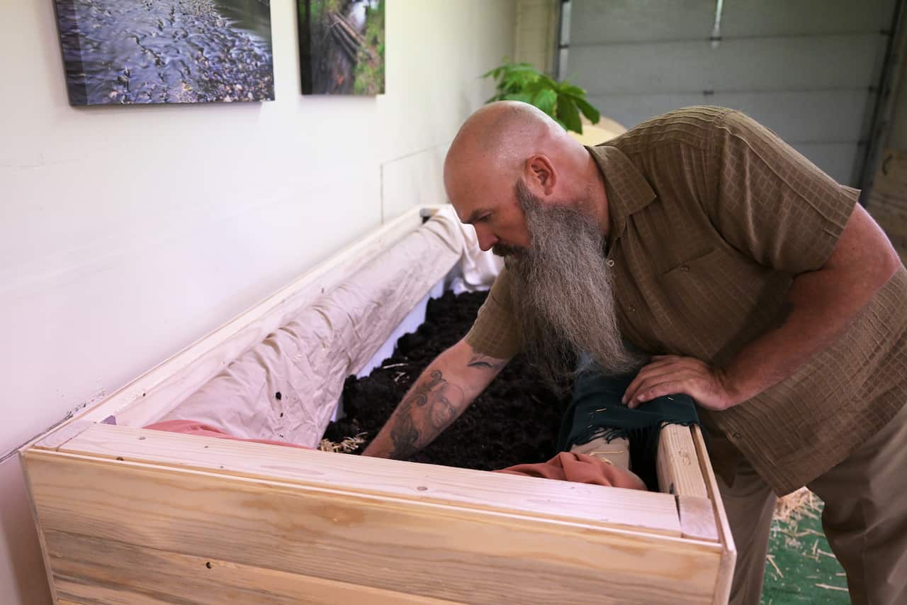 A man with a long grey beard reaching into an open wooden box containing earth, inside a room.