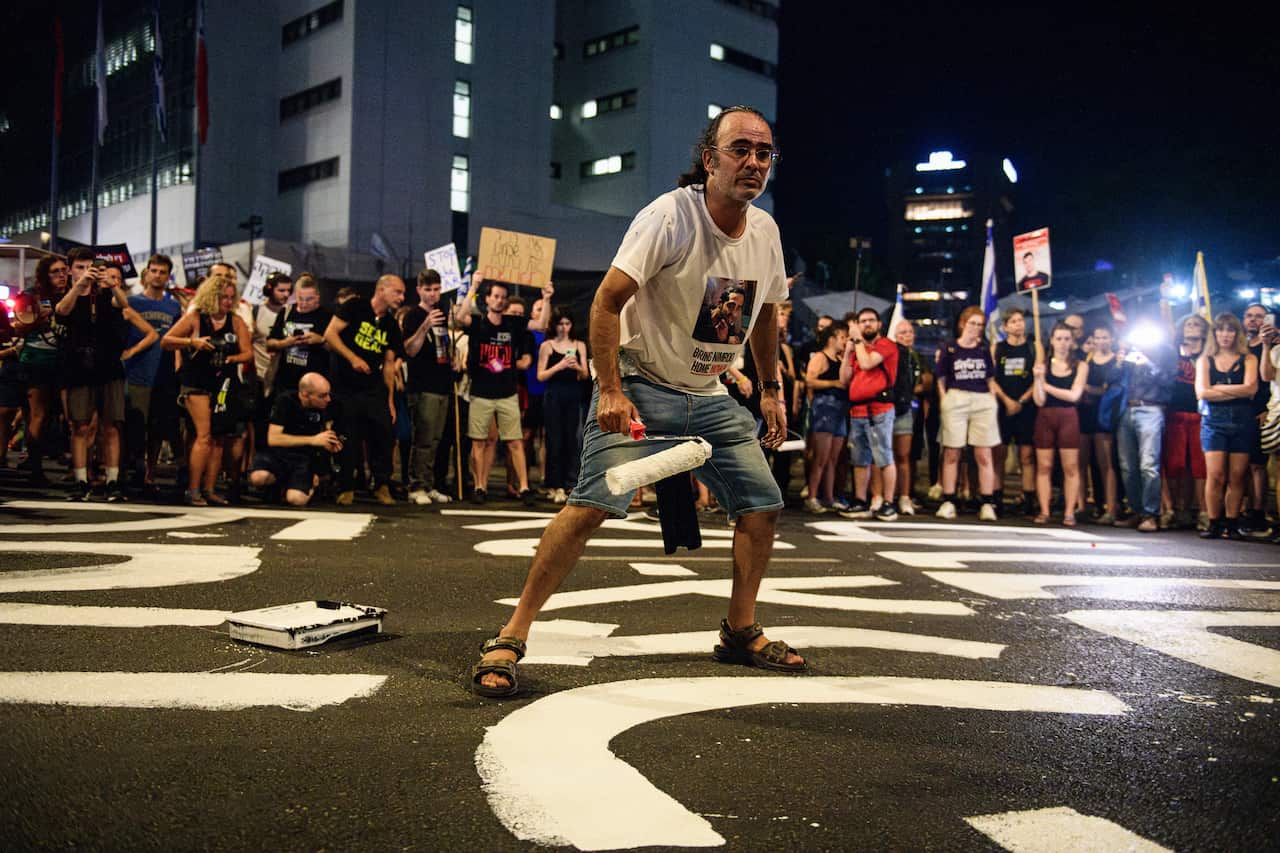  A man wearing jean shorts and a white t-shirt paints a slogan on the road as part of a protest. Other protesters stand further behind him, holding signs. 