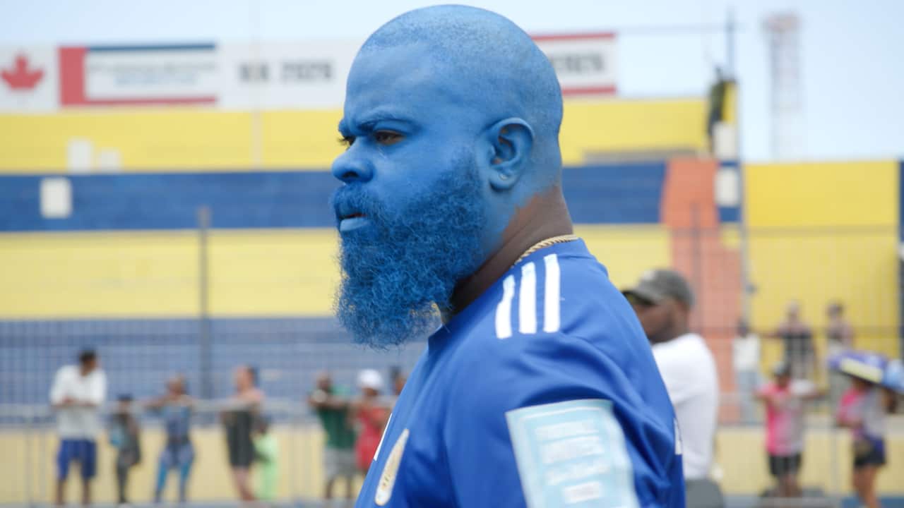 A man with a beard wears a blue football jersey. His head and face are painted blue.