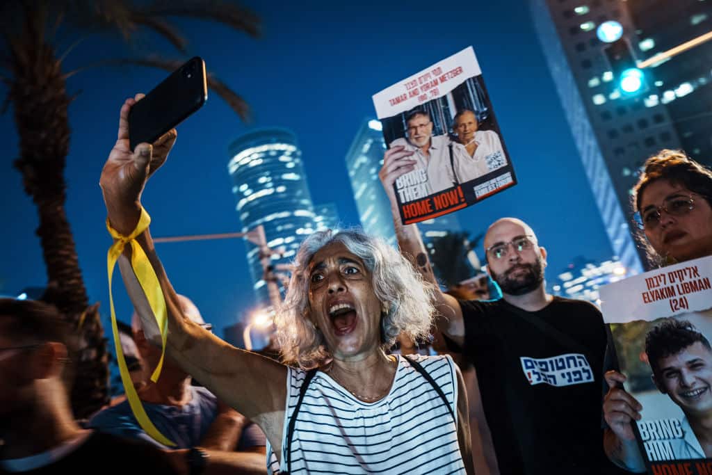A woman in a striped shirt holds up a phone. Her mouth is open, as if she is shouting. People beside and behind her hold up photos of Israelis held hostage by Hamas.