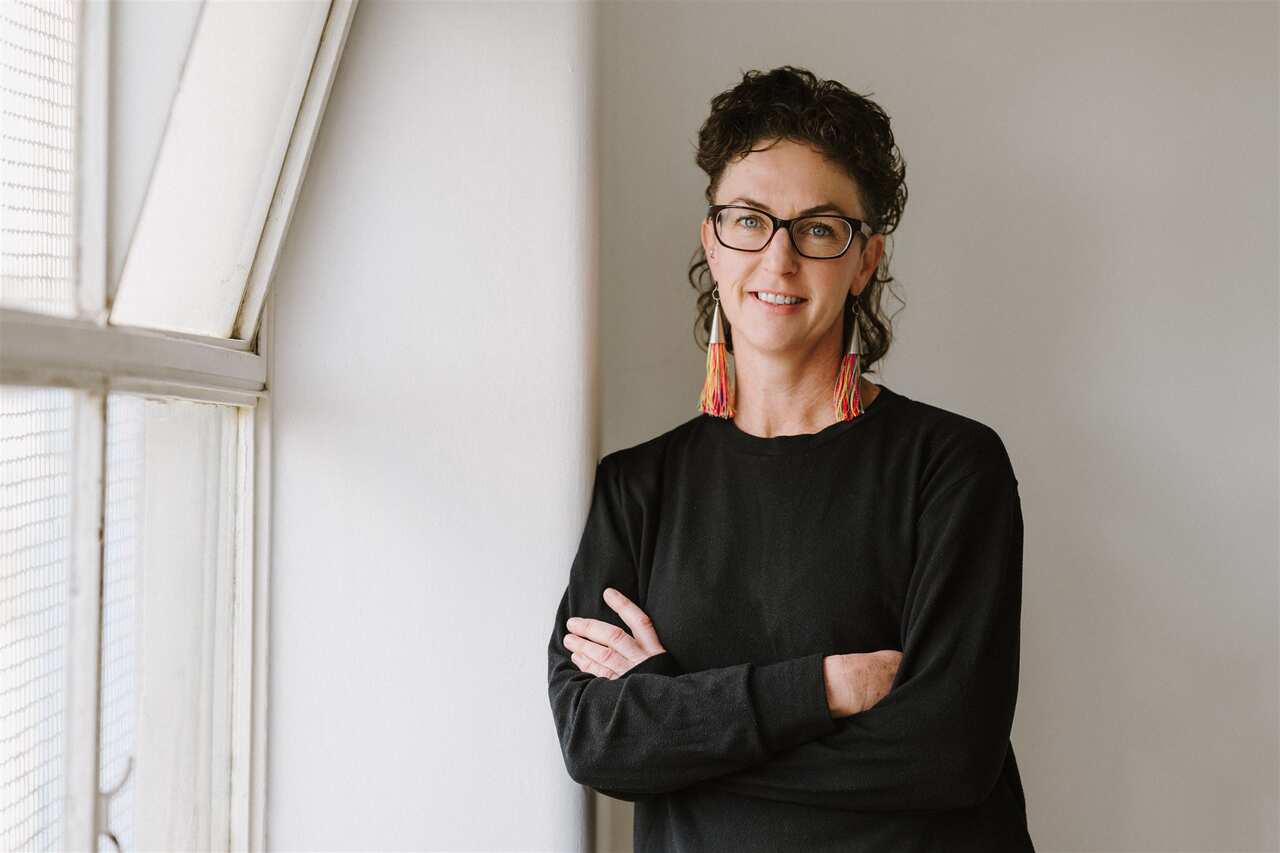A woman with glasses and long, colourful tassel earrings, wearing a long-sleeved black shirt, stands with her arms crossed next to a window.
