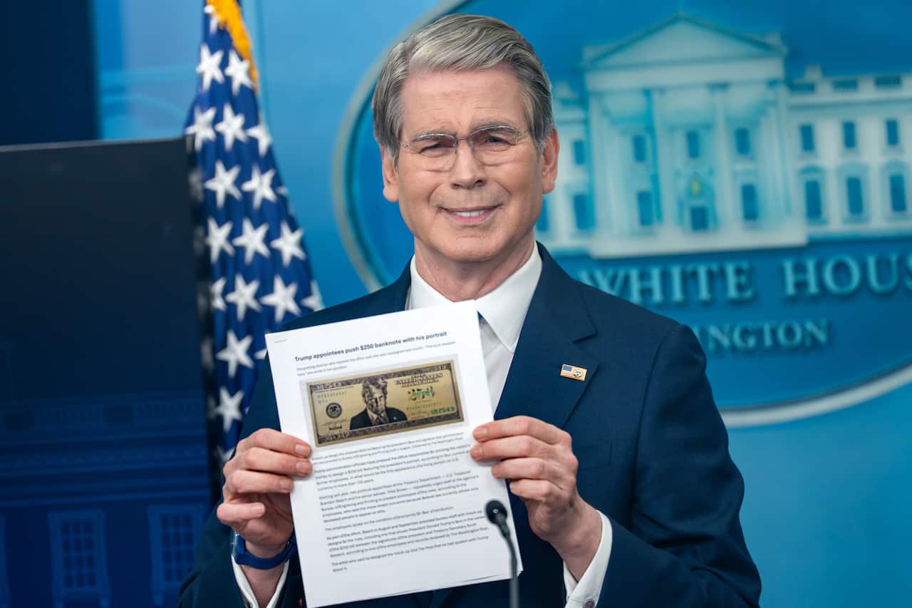 A man in a dark suit stands at a White House briefing room podium, holding up a printed page that includes an image of a mock banknote featuring Donald Trump's portrait. An American flag is visible in the background alongside a blurred White House emblem.