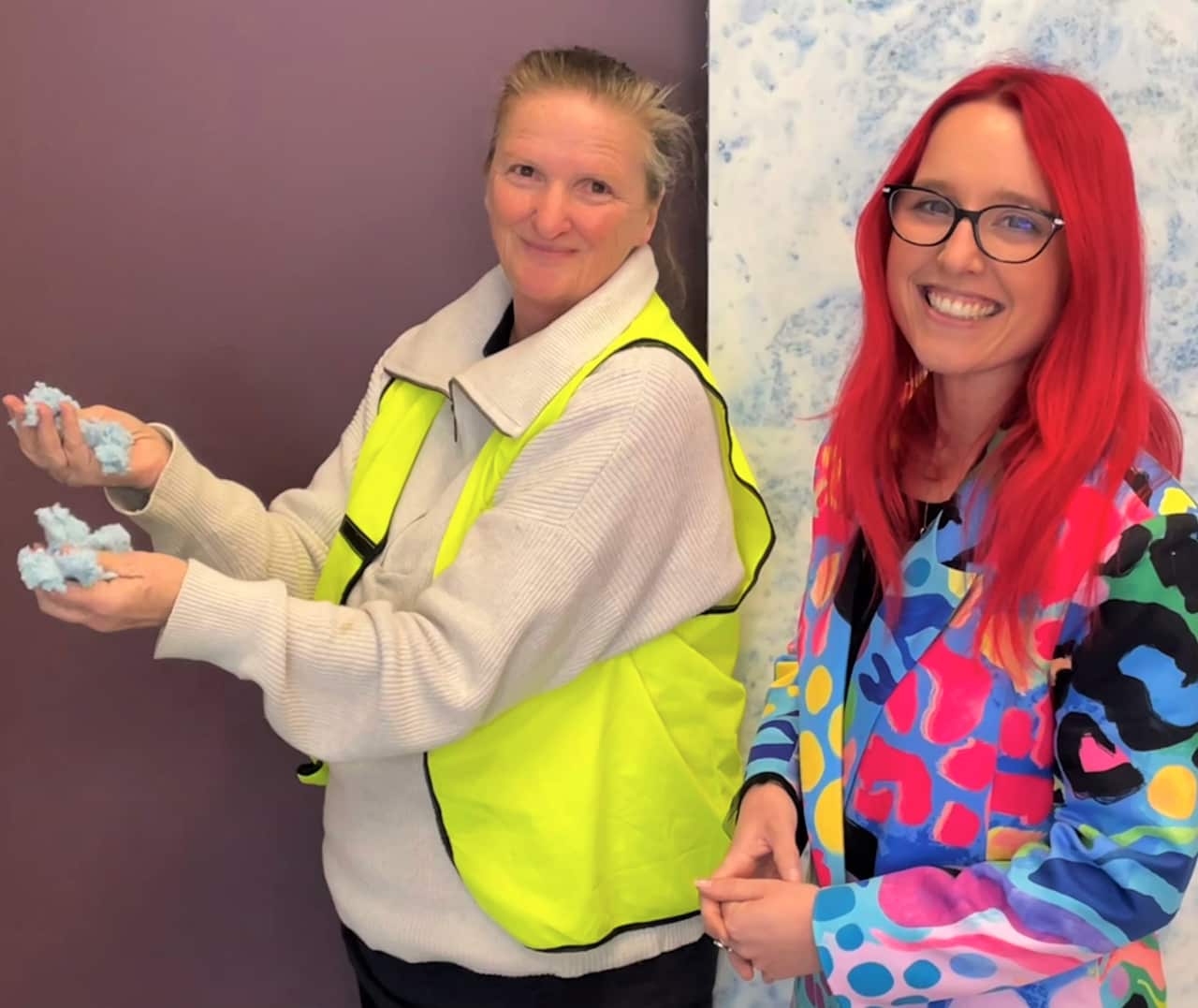 A woman in a hi-viz vest holds shredded fabric in her hands while another woman with pink hair and a multi-coloured jacket looks at camera. 