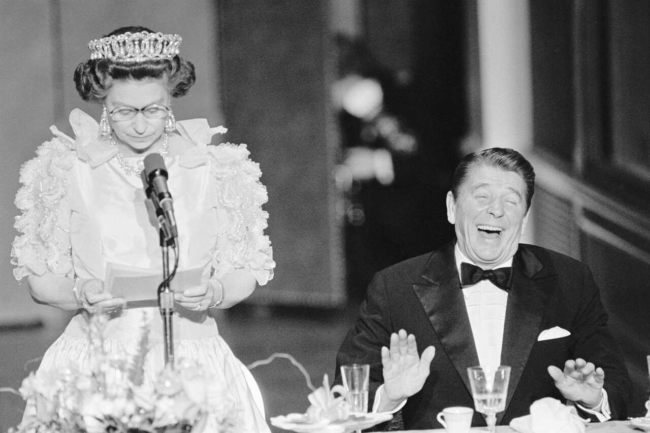 Queen Elizabeth II (left) stands and speaks in front of a microphone while then US President Ronald Reagan, seated next to her, laughs.