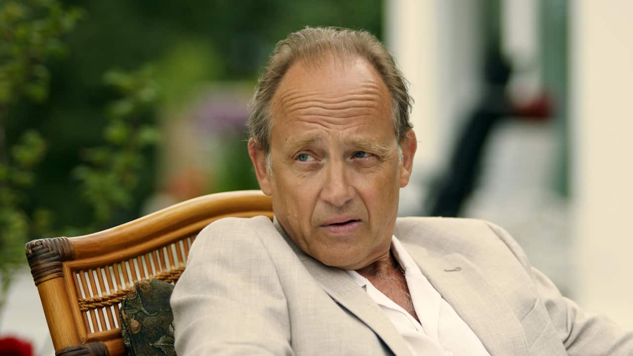 A man in a pale cream suit sits in a wicker chair, looking sideways. 