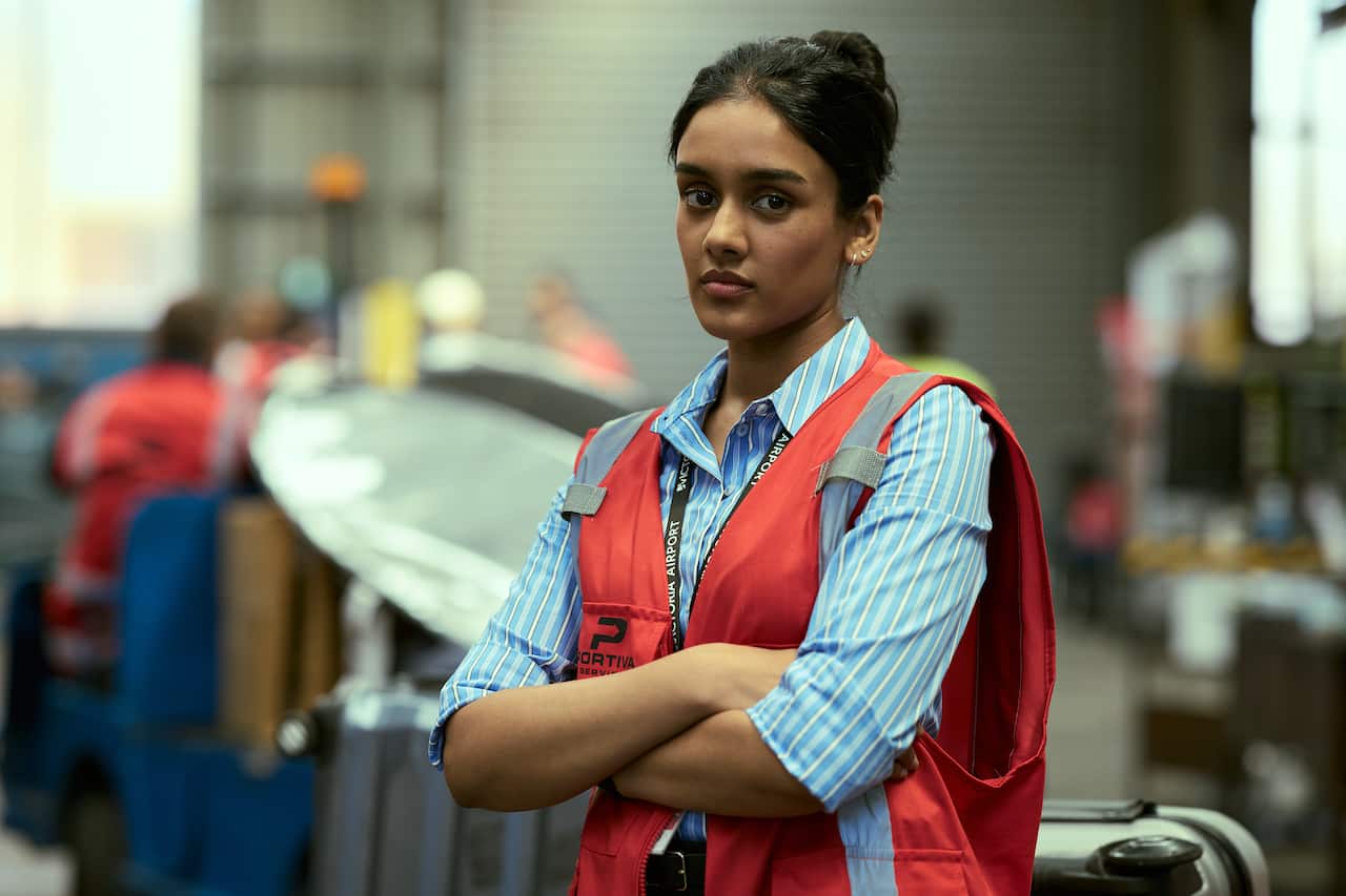 A woman in a collared blue shirt with a red vest over it stands with her hands clasped across her chest.