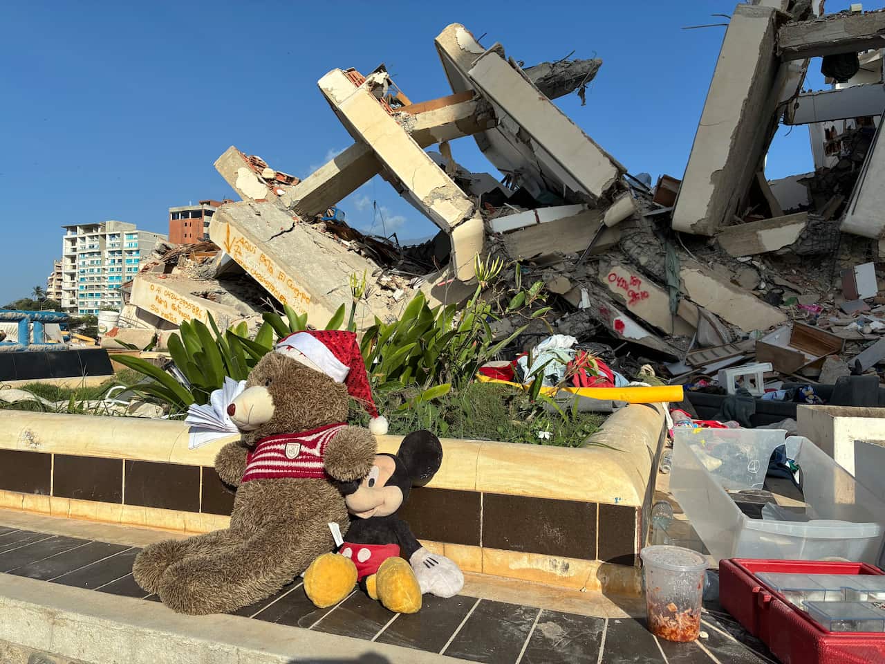 Two stuffed toys, a brown bear wearing a Santa hat and a Mickey Mouse, sit in front of a building destroyed by an earthquake.