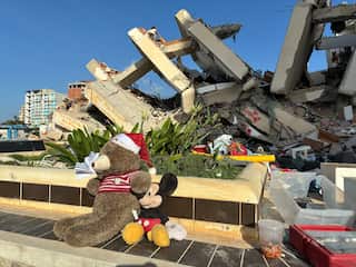 Two stuffed toys, a brown bear wearing a Santa hat and a Mickey Mouse, sit in front of a building destroyed by an earthquake.