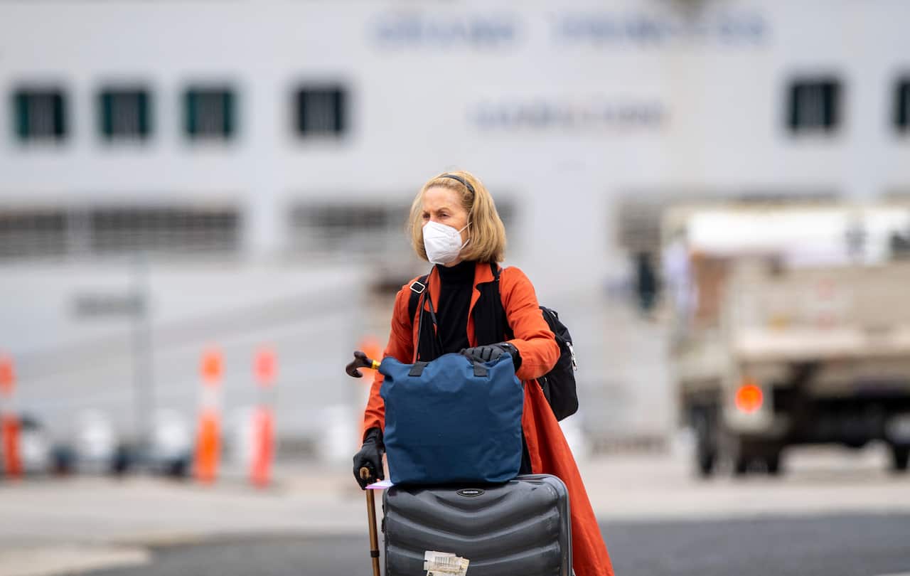 A woman wearing a face mask in an orange coat with a blue bag on top of a navy suitcase.