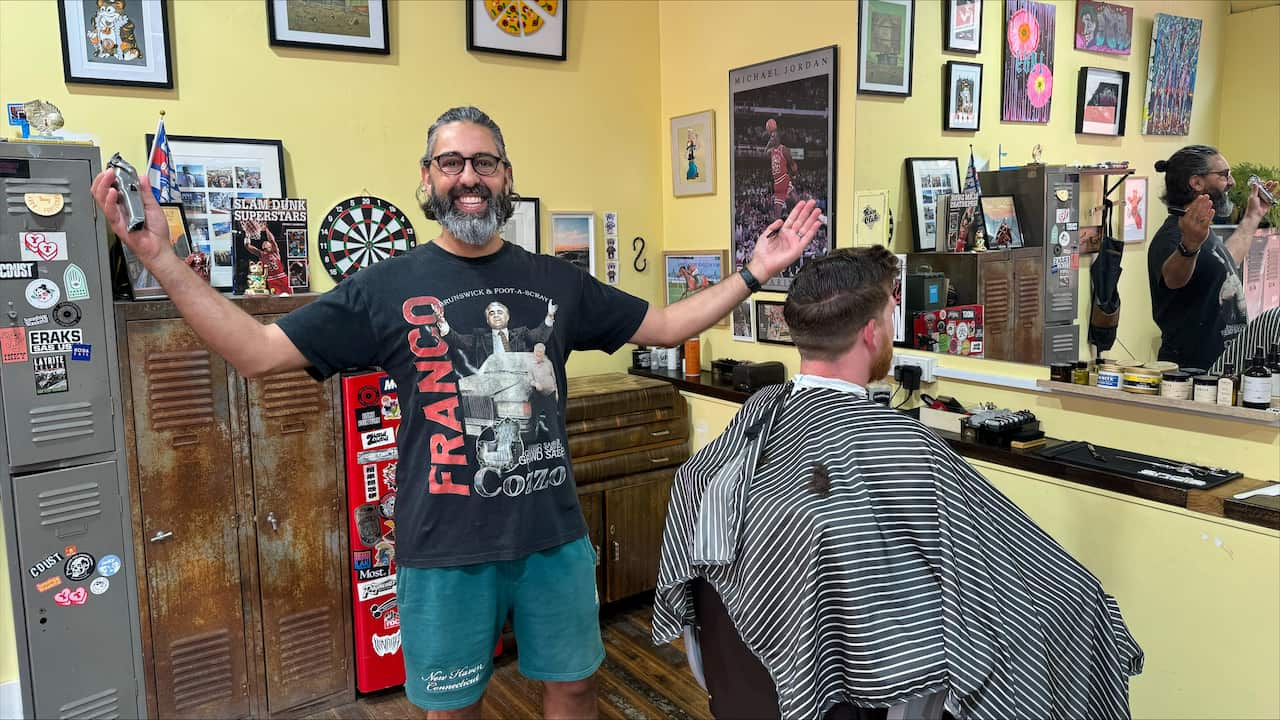 A male hairdresser wearing glasses and a black T-shirt stands with his arms spread wide and a smile on his face.