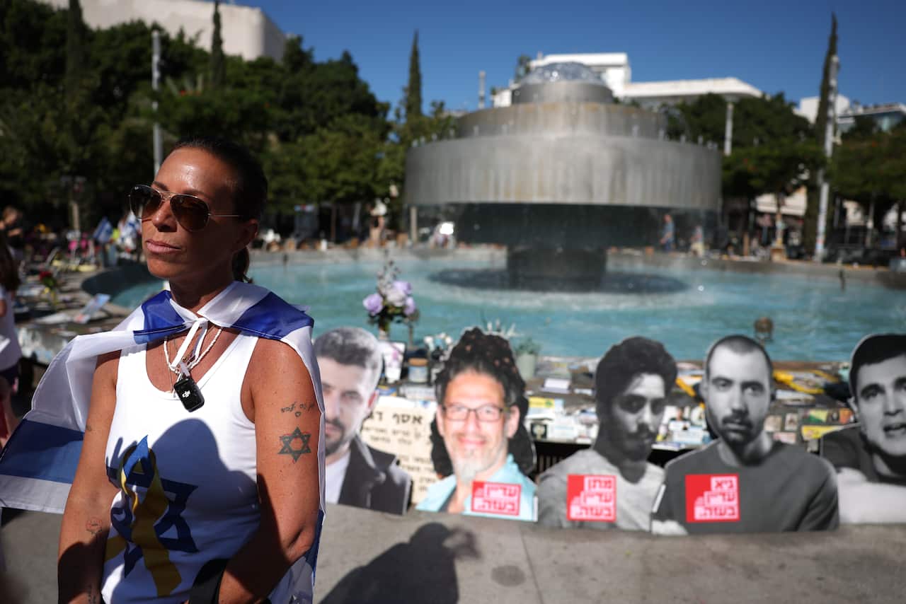 A woman wearing an Israeli flag on her back stands in front of a fountain and images of October 7 victims that have been placed there.