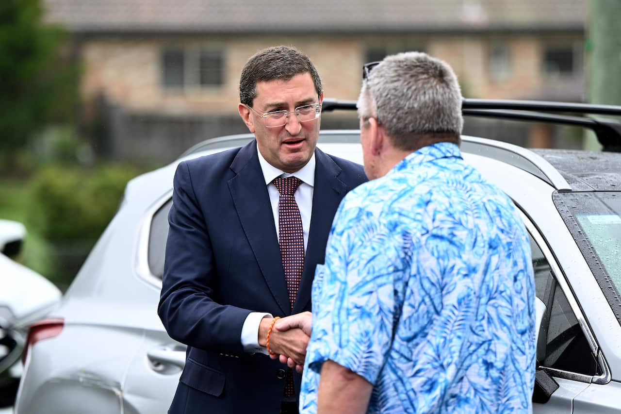 Two men, one in a blue suit and the other in a blue floral print shirt are shaking hands beside a parked car.