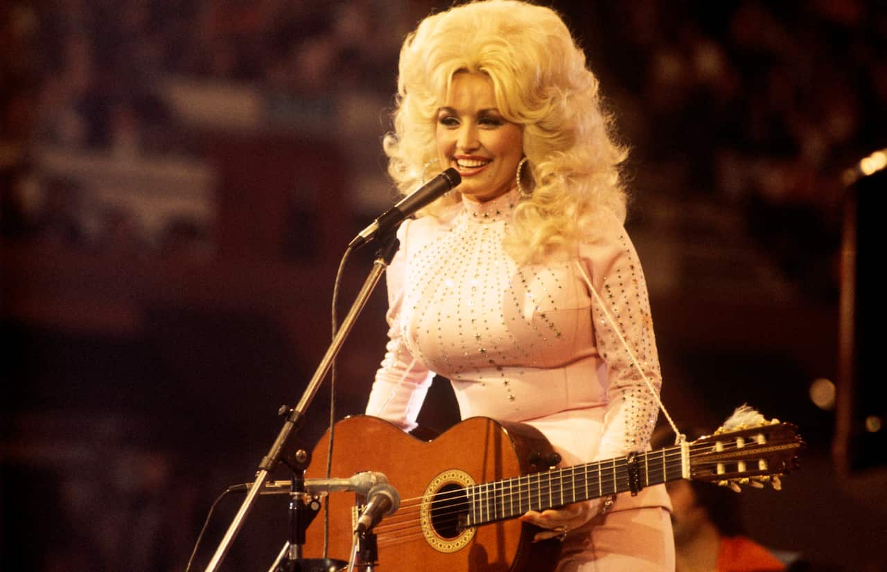 Person standing at a microphone with an acoustic guitar, wearing a light-coloured embellished outfit during a live stage performance.