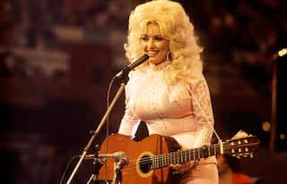 Person standing at a microphone with an acoustic guitar, wearing a light-coloured embellished outfit during a live stage performance.