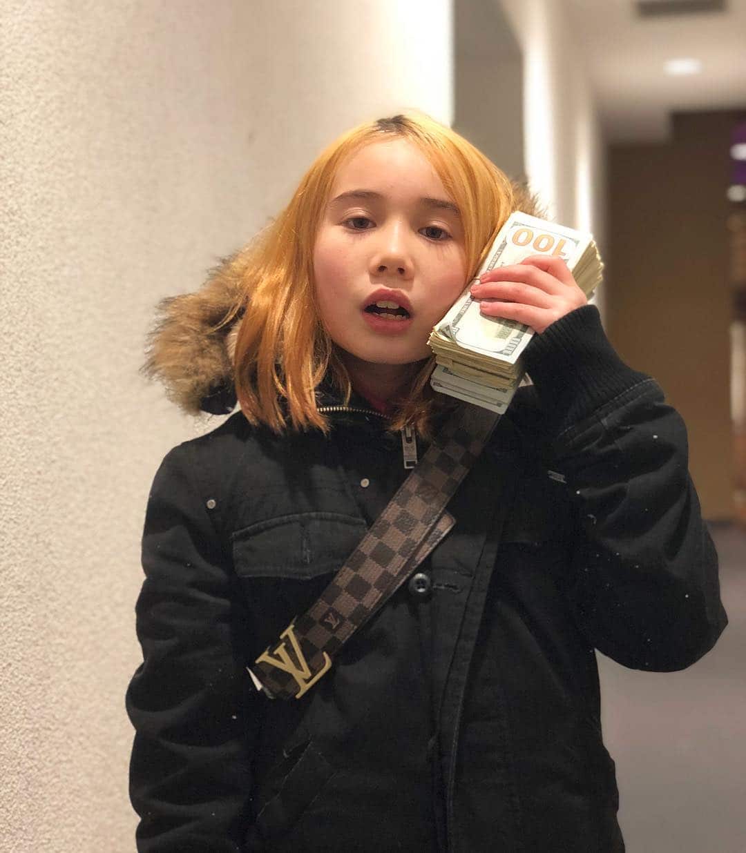 A young girl wearing a black jacket with a Louis Vuitton bag over her shoulder, holding a wad of cash against her face.