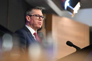 A man with glasses and a red tie stands at a podium speaking into a microphone.