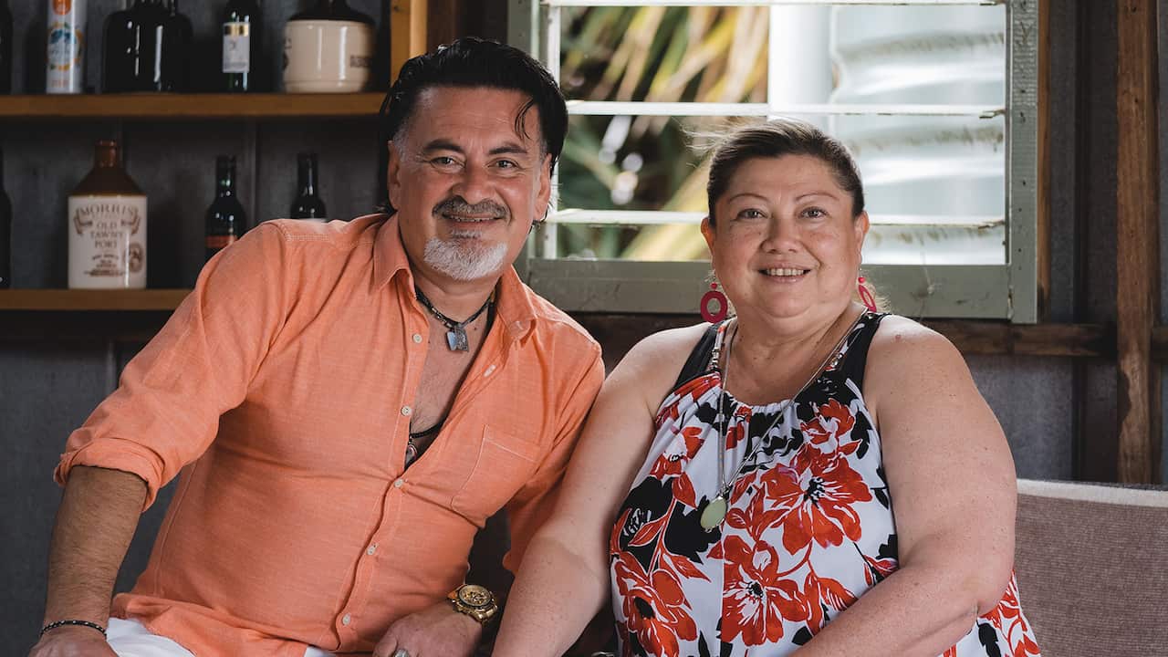 A man in an orange shirt and a woman in a floral top sit together, smiling. 