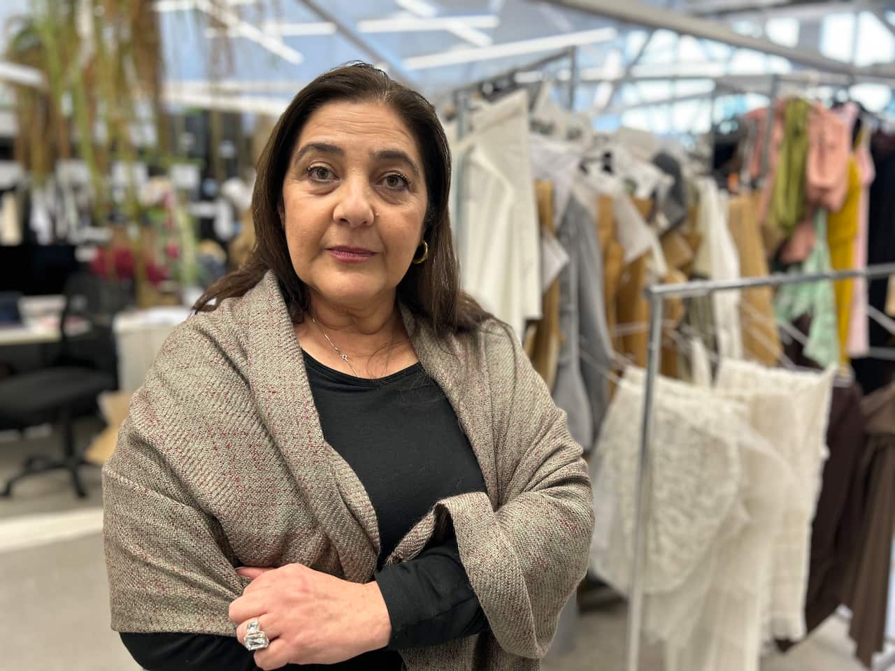 A woman with dark hair, a black top and grey shawl stands in a warehouse. Behind her are clothes hanging on racks.