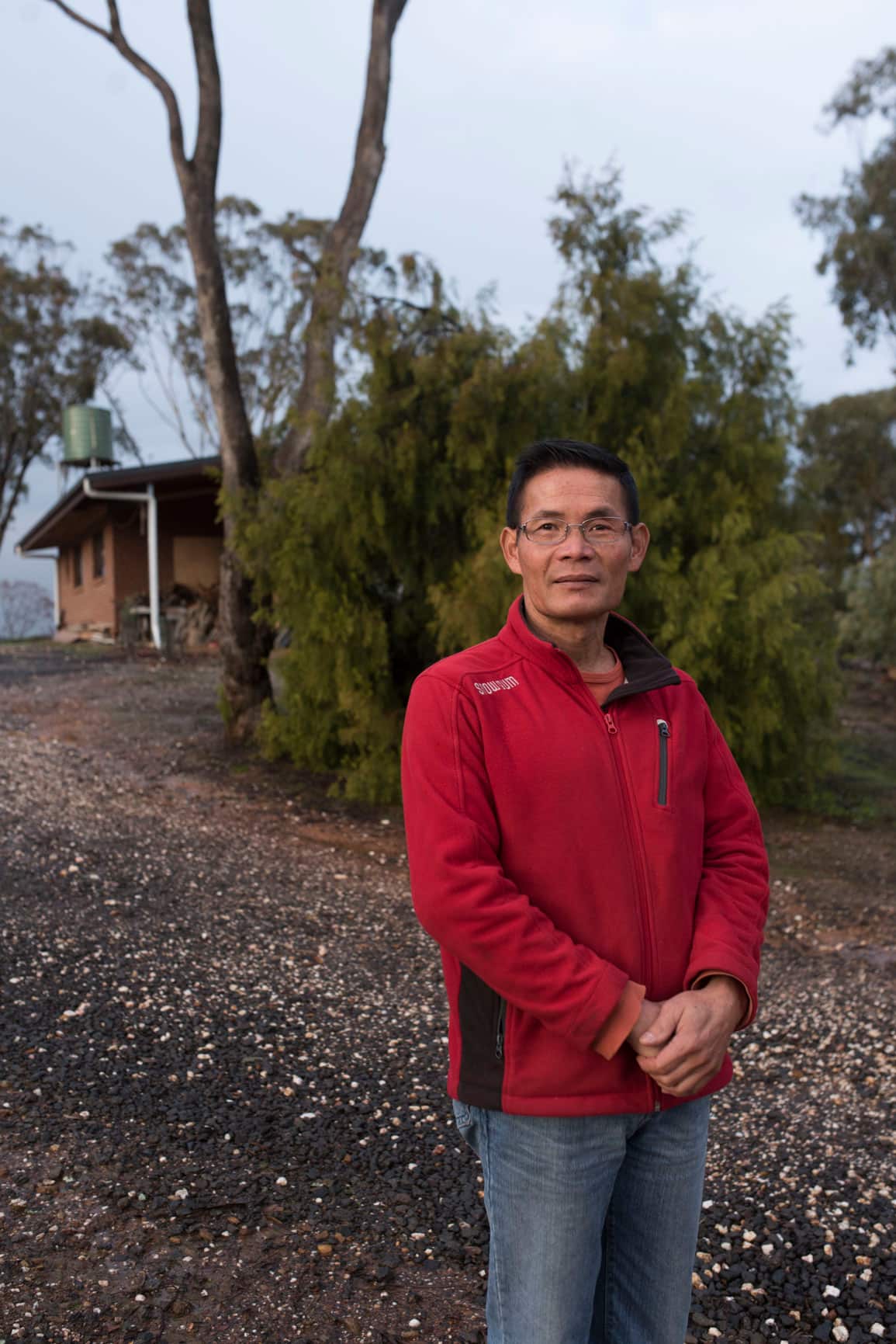 A man wearing a red fleece and blue jeans stands in front of a tree and brick building 