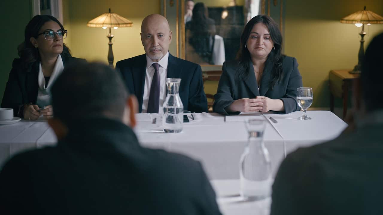 A medium shot shows three people in professional attire sitting behind a long white-clothed table in a dimly lit room, facing two individuals in the foreground whose backs are to the camera.