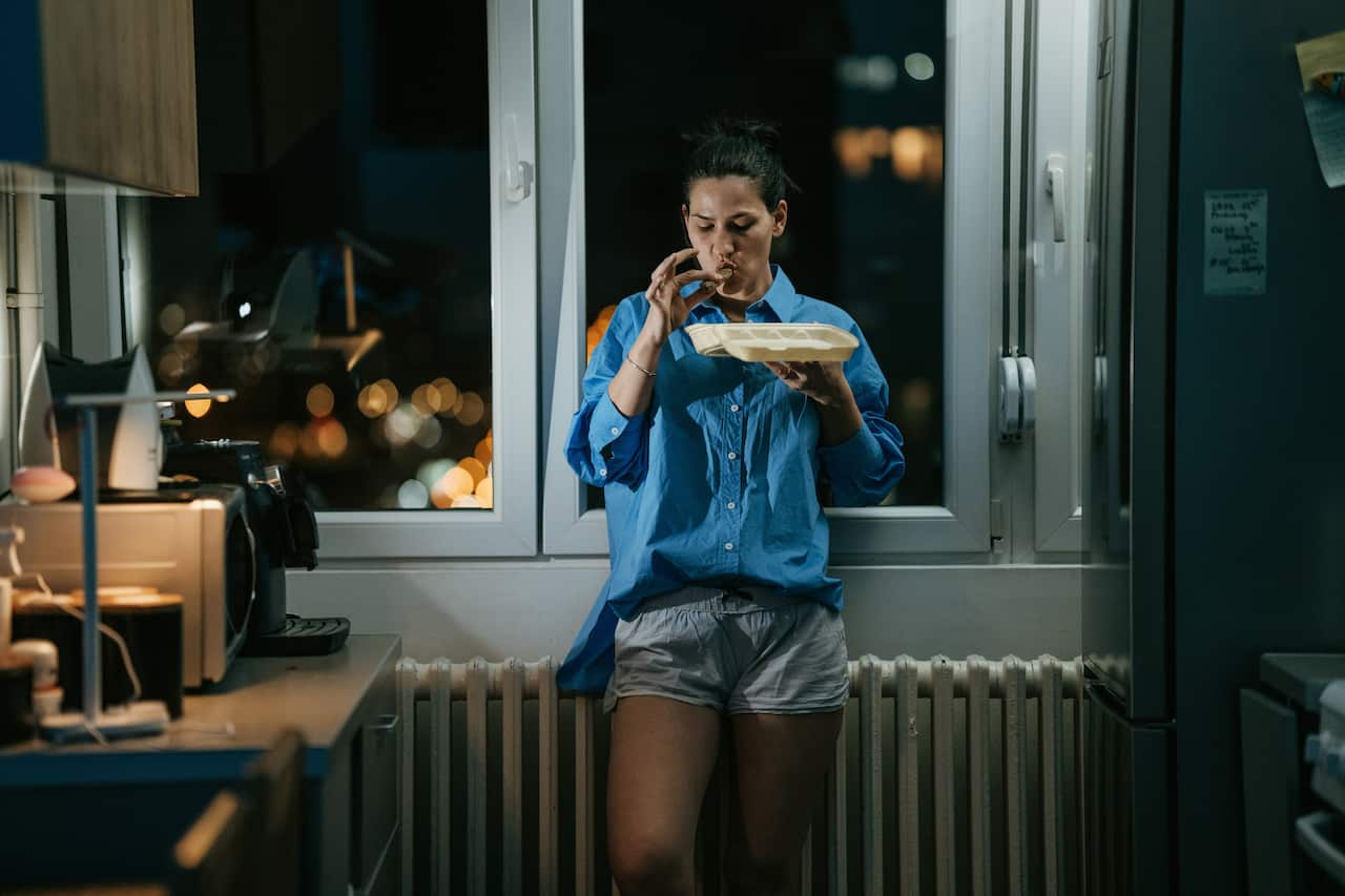 Woman standing in kitchen late at night eating food.