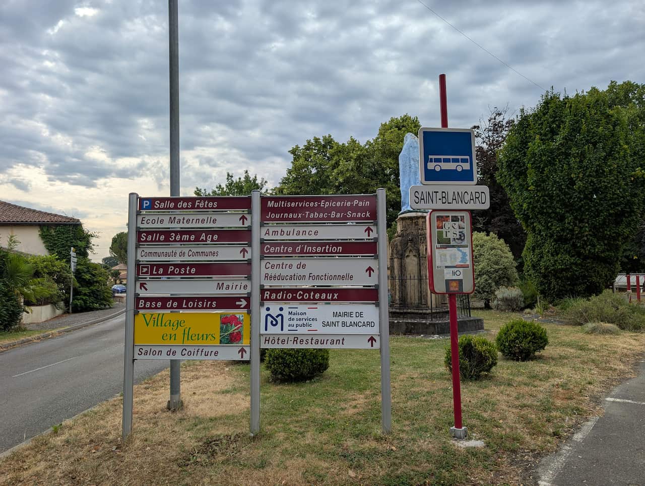Two sets of road signs in French, as well as a bus stop with a sign read 'Saint Blancard'. Trees and a statue are visible in the background.