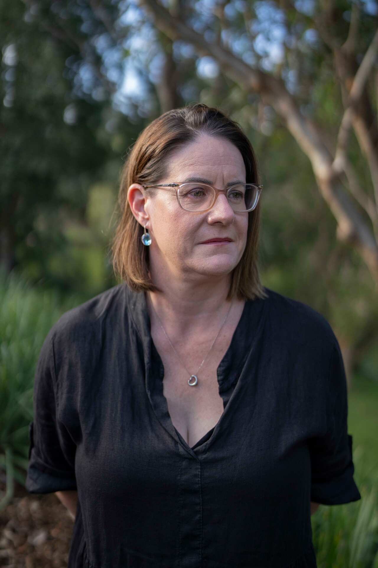 A middle-aged woman with shoulder-length brown hair wears a black blouse and glasses and is standing in a park.