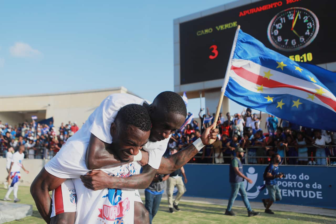 A jubilant football player perched on a teammate's shoulders, who is holding the Cape Verde flag in one hand as they celebrate their team's victory in a stadium.