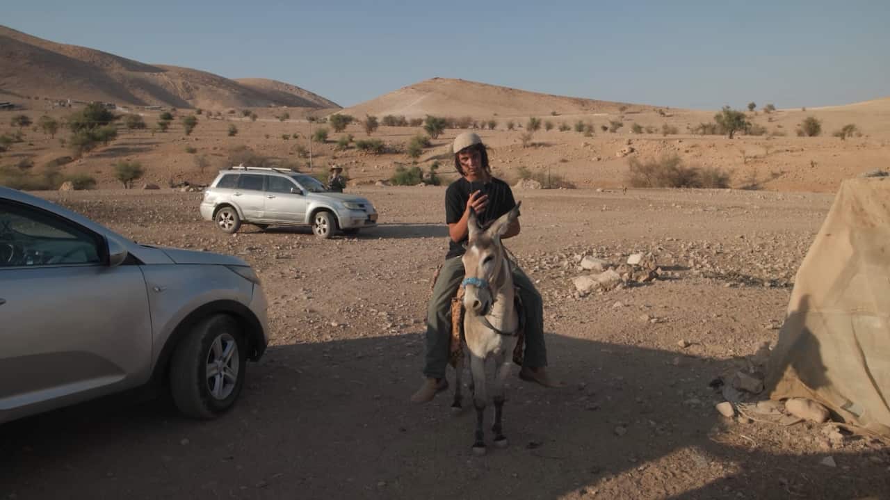 A teenager holding up a phone sits on top of a donkey. He is in a dry, rocky landscape and there are two cars behind him. 